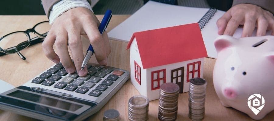 Person calculating finances with a model house, coins, and piggy bank.