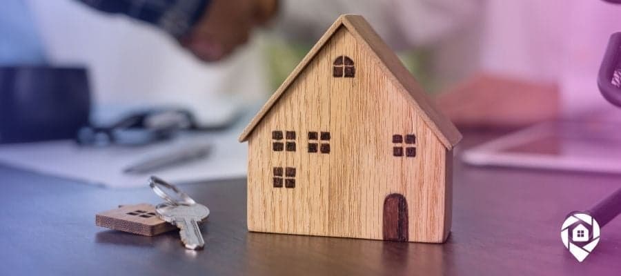A wooden house on a table with a key next to it and people shaking hands in the background for seller broker vs listing broker.
